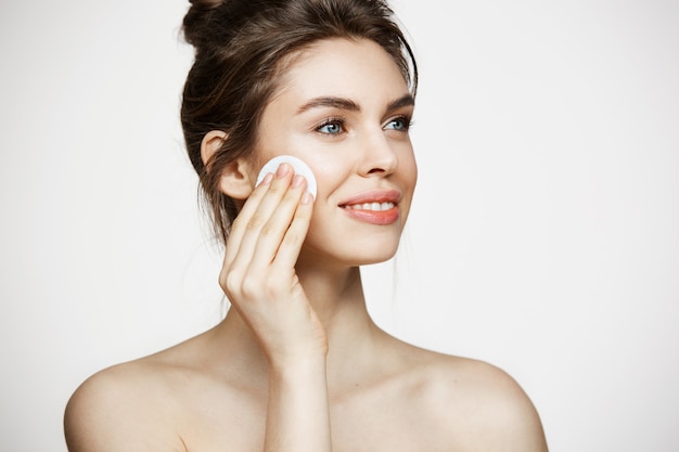 Beautiful natural brunette girl cleaning face with cotton sponge smiling over white background. Cosmetology and spa.