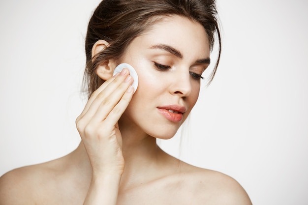 Beautiful natural brunette girl cleaning face with cotton sponge smiling over white background. Cosmetology and spa.