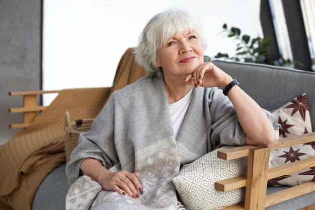 Beautiful neat sixty year old grandmother wearing wide gray scarf and wrist watch resting comfortably on couch in living room, smiling happily, waiting for her son and grandchildren to come