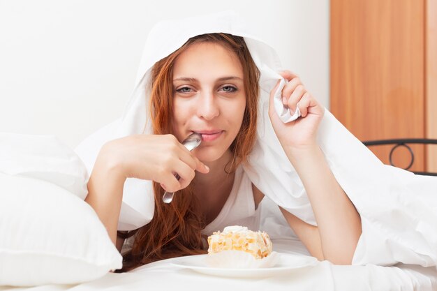 Beautiful ong-haired woman eating sweet cake under sheet in bed