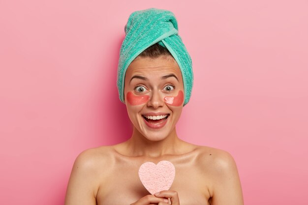 Beautiful overjoyed woman wears hydrating eye patches, smiles happily, shows white teeth, holds cosmetic sponge, has bare shoulders, models against pink background.