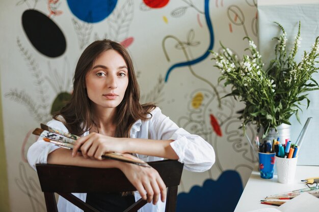 Beautiful painter in white shirt on chair dreamily looking in camera while holding paint brushes in hand at home