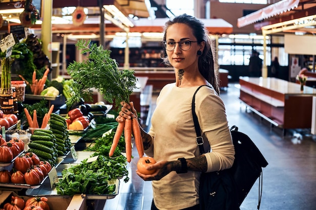 Beautiful pensive girl in glasses is buying fresh carrots at local farmer's market.