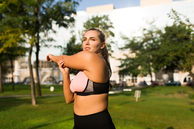 Beautiful pensive plus size girl in pink sporty top and leggings thoughtfully looking aside while stretching in city park