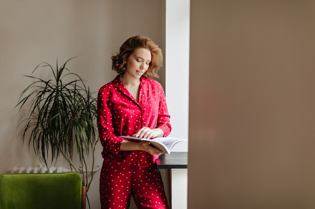Beautiful pensive woman reading magazine in morning. Indoor shot of attractive woman in red sleepwear.