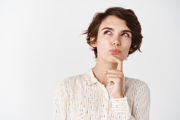 Foto gratuita bella donna pensierosa che tocca il mento e alza lo sguardo, fa una scelta o pensa, in piedi contro il muro bianco