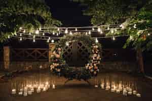 Free photo beautiful photozone with big wreath decorated with greenery and roses in centerpiece, candles on the sides, and garland hanged between trees