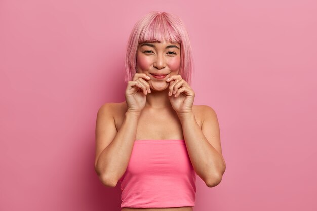 Красивая розоволосая азиатка приятно улыбается, держит руки у губ, радуясь приятным новостям, носит розовые топы в помещении. Монохромный. Этническая девушка с модной стрижкой выражает хорошие эмоции