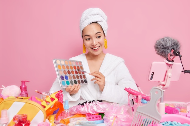 Beautiful pleased Asian woman holds eye shadow palette going to do make up talks with followers 