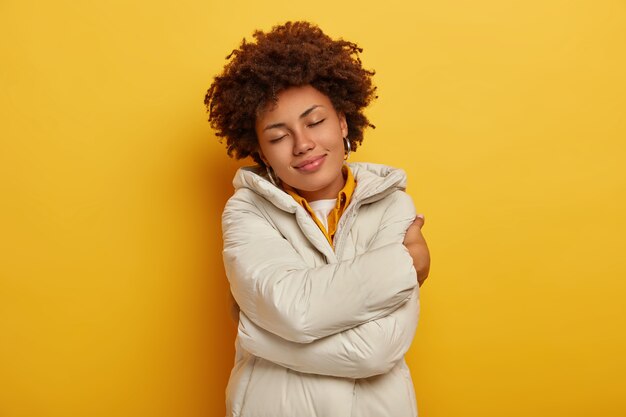 Beautiful pleased woman enjoys comfort in new winter jacket, embraces herself, keeps eyes shut, feels warm and satisfied, har curly hairstyle, isolated over yellow background. People, clothes concept