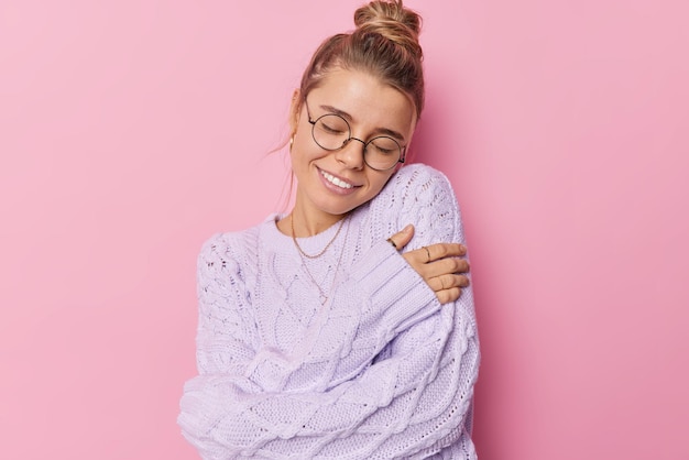 Beautiful pleased young woman with fair hair gathered in bun embraces own body wears knitted sweater expresses self love takes care of herself isolated over pink background Self acceptance