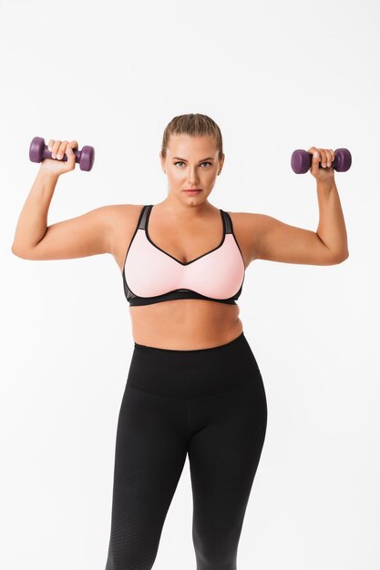 Beautiful plump girl in sporty top and leggings holding dumbbells in hands while thoughtfully looking in camera over white background