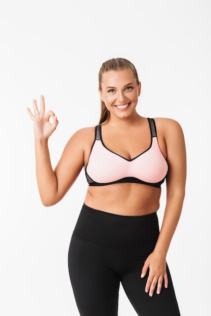 Beautiful plump girl in sporty top and leggings joyfully showing ok gesture while looking in camera over white background