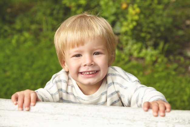 Beautiful portrait of blond smiling child. Little kid playing outside in summer garden near home. Brown eyes, milk teeth, tiny fingers are incredibly nice. Childhood concept.