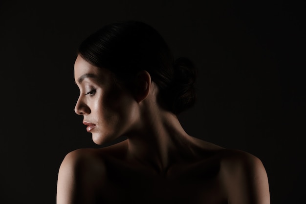 Beautiful portrait of half-naked elegant woman with dark hair in bun putting head aside, isolated over black