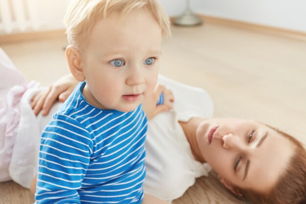 Beautiful portrait of little blond boy with blue eyes and caring mother lying on the floor at home. Tiny baby in blue clothes looking ahead. His attractive careful mom watching him with love.