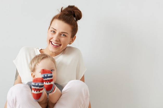 Beautiful portrait of smiling mother and a child sitting together on the white wall. Happy European woman in white clothes smiling and holding her son in colorful socks on her legs.