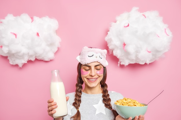 Beautiful positive woman with two pigtails smiles gladfully looks at bowl of cornflakes