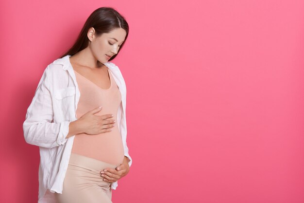 Beautiful pregnant woman hugging her tummy against pink space, looking at her belly with love, copy space for advertisement or promotional text text.