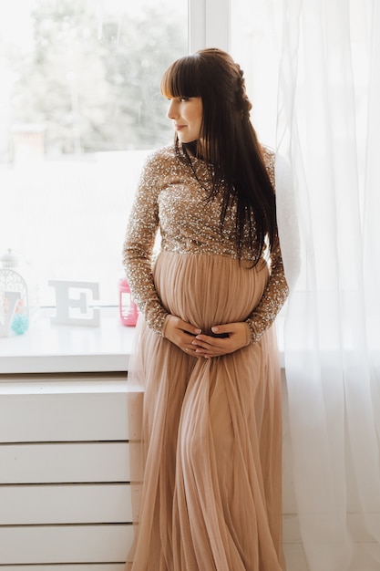 Красивая беременная женщина в длинном бежевом платье, стоя в яркой уютной комнате