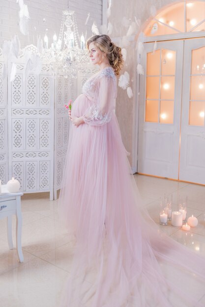 豊かなピンクのドレスの美しい妊娠中の女性は、彼女の腹のポーズで手を保持しています