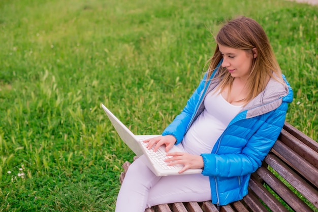 Free photo beautiful pregnant woman sitting on bench using laptop outside.