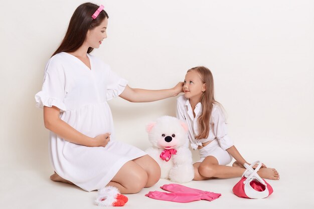 Красивая беременная женщина сидит с дочерью на полу в окружении детской одежды и мягкой игрушки