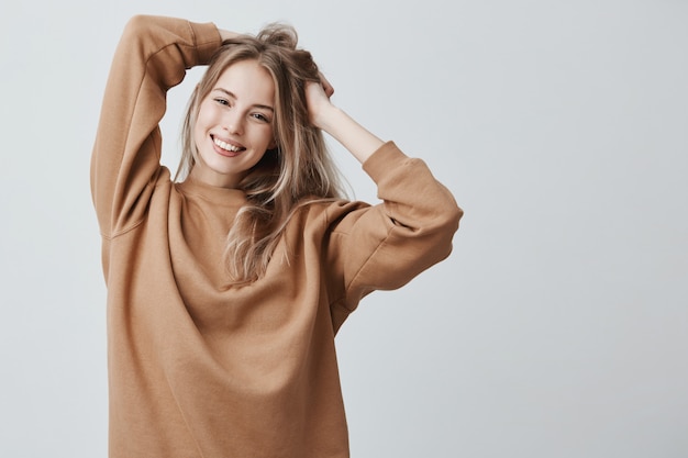 Beautiful pretty charming young blonde woman in loose sweater smiling happily, having fun indoors, playing with long straight hair.