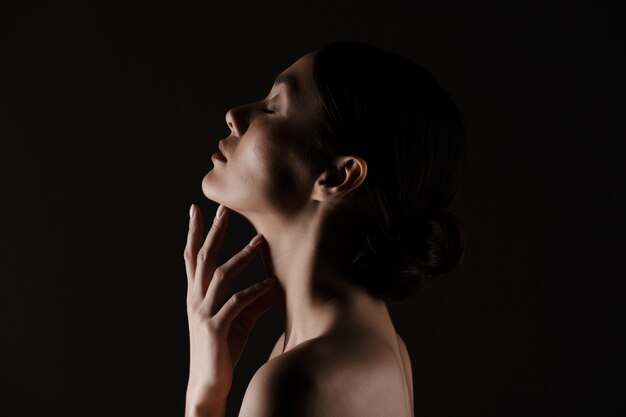 Beautiful in profile of half-naked gentle woman posing on camera with closed eyes isolated, over black