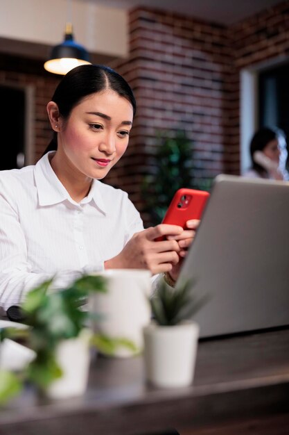 Beautiful project manager in office workspace with smartphone device sitting at desk while texting workplace colleague. Company employee using touchscreen phone to send messages at work.