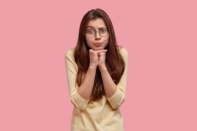 Beautiful puzzled female model keeps hands together under chin, listens attentively information, has curious worried expression, dressed in casual jumper, isolated over pink wall. Human emotions