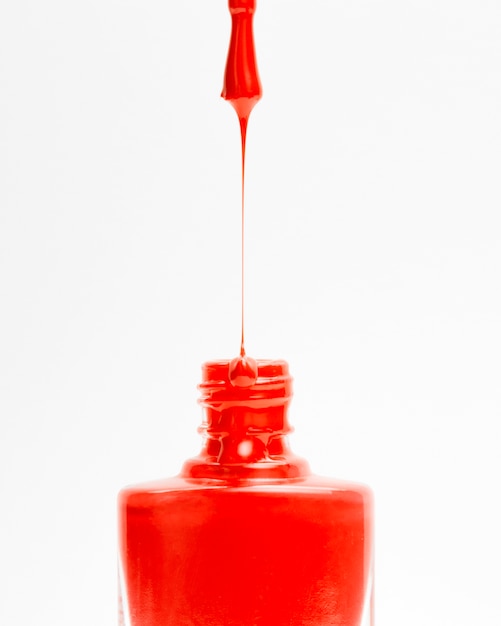 Beautiful red colored nail polish dripping from brush into bottle on white backdrop