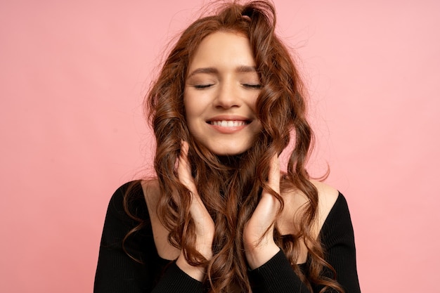 Beautiful red head woman with close eyes posing over pink wall. Wavy hairs. Perfect smile. 