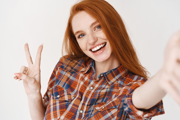 Красивая рыжая девушка счастливо улыбается и показывает знак мира, делая селфи на передней камере смартфона на белом фоне