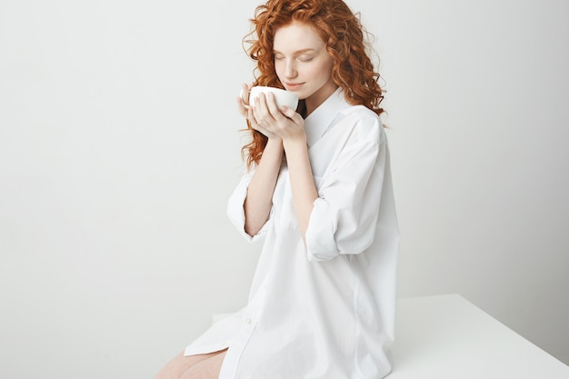 Beautiful redhead girl with curly hair smiling holding cup sitting on table . Closed eyes.
