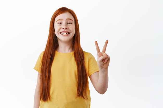 Beautiful redhead girl with freckles smiling broadly, showing two fingers peace sign and looking happy, standing in yellow t-shirt against white background