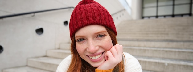 Free photo beautiful redhead student girl in red hat smiles sincere looks happy and relaxed sits on stairs