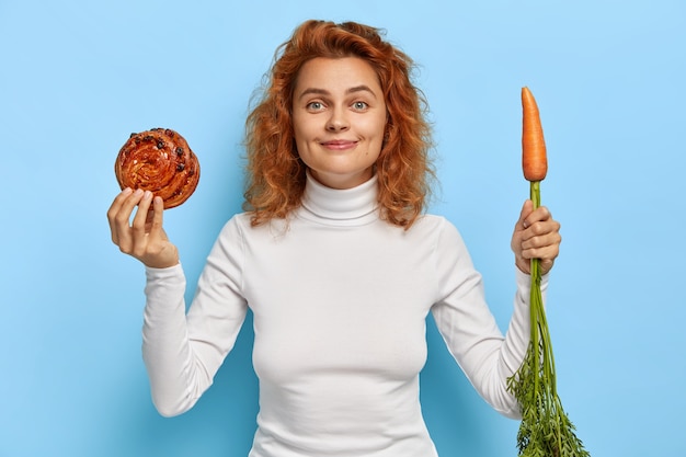 Beautiful redhead woman chooses between fresh carrot and sweet bun