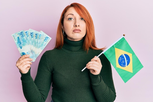 Beautiful redhead woman holding 100 brazilian real banknotes and brazil flag relaxed with serious expression on face. simple and natural looking at the camera.