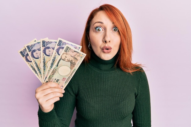 Beautiful redhead woman holding japanese yen banknotes scared and amazed with open mouth for surprise, disbelief face