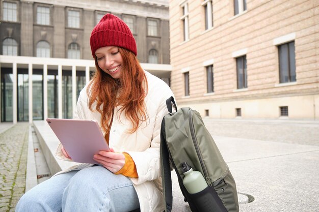 Beautiful redhead woman in red hat sits with backpack and thermos using digital tablet outdoors conn