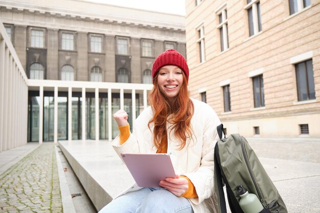 Beautiful redhead woman in red hat sits with backpack and thermos using digital tablet outdoors conn