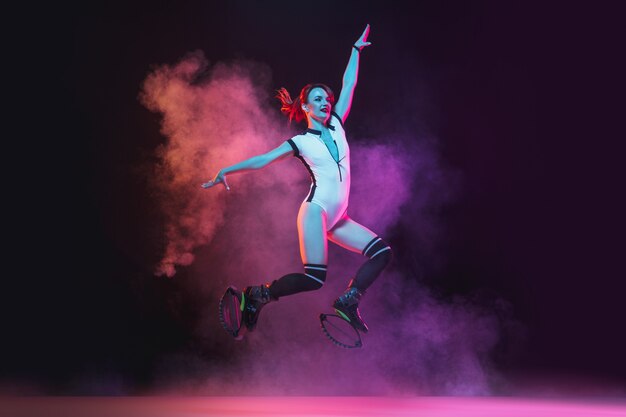 Beautiful redhead woman in a red sportswear jumping in a kangoo jumps shoes on dark background.