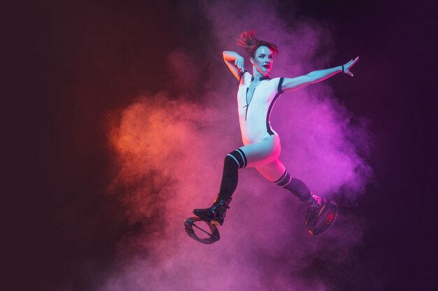 Beautiful redhead woman in a red sportswear jumping in a kangoo jumps shoes on dark studio background.