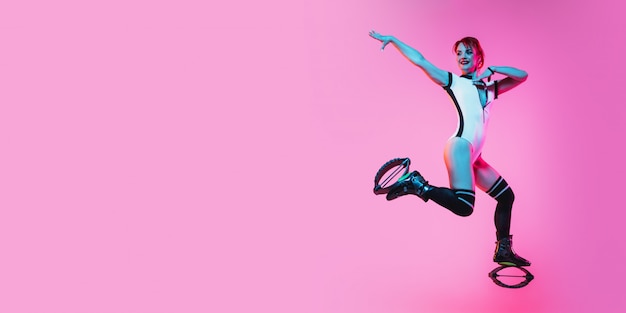 Beautiful redhead woman in a red sportswear jumping in a kangoo jumps shoes on pink studio background. Flyer