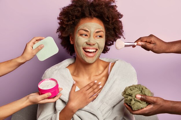Beautiful refreshed cheerful woman with nourishing clay mask looks happily aside, being treated by cream, sponges and makeup brush