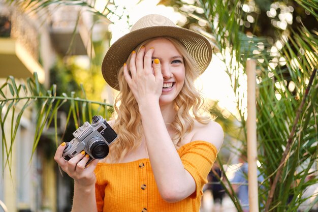 Beautiful romantic blond girl in hat happily holding retro camera in hand on city street
