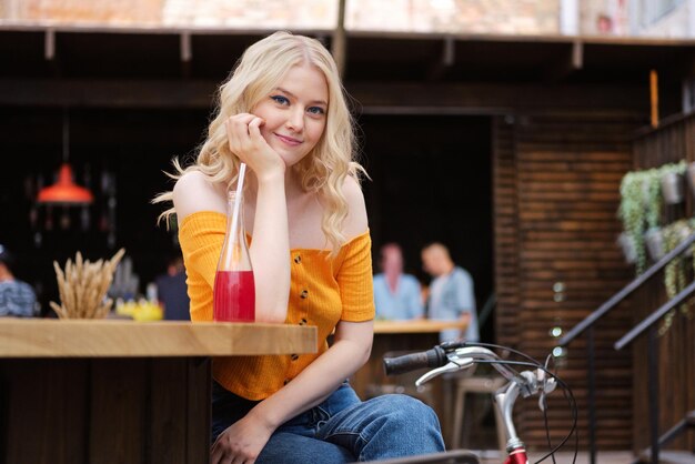 Beautiful romantic blond girl leaning on hand dreamily looking in camera with lemonade in courtyard of city cafe