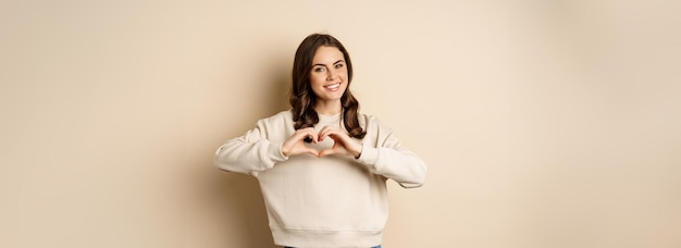 Foto gratuita la bella donna castana romantica che mostra il segno di amore del cuore si prende cura di te che sorride alla macchina fotografica che sta ov