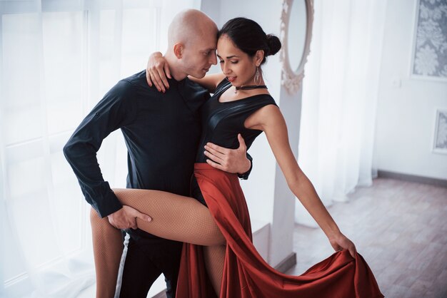 Beautiful romantic pose. Young pretty woman in red and black clothes dancing with bald guy in the white room